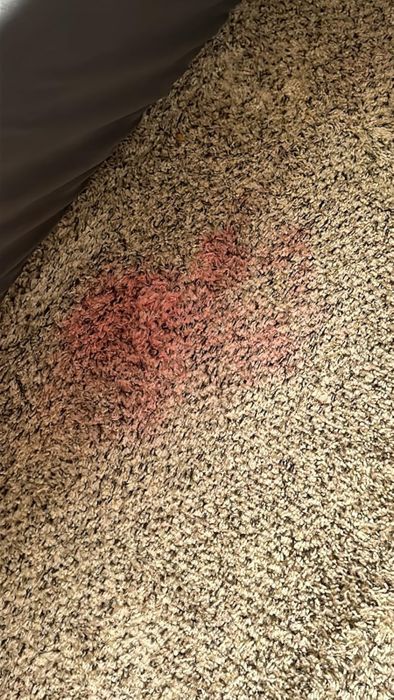 A red stain on a carpet near a piece of furniture's corner.