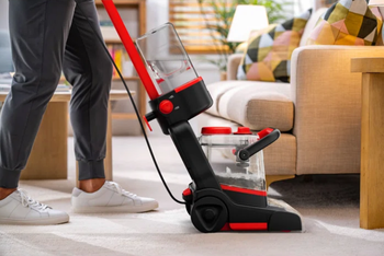 A person operating a carpet cleaner in a modern living room with stylish furniture. The cleaner’s canister is visible with soapy water inside.