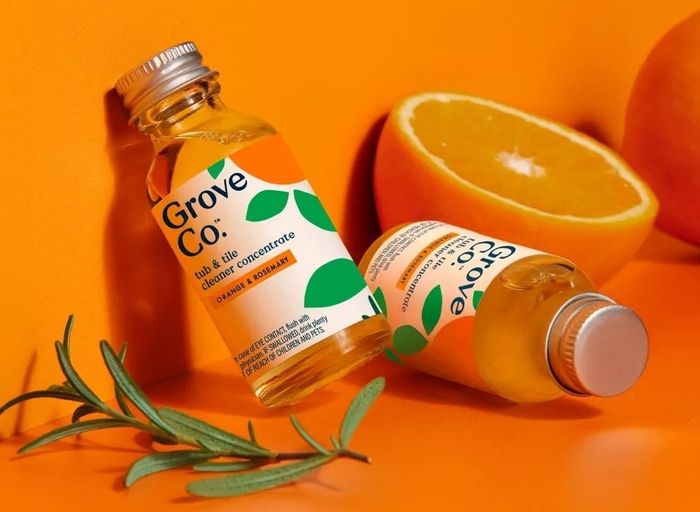 Two bottles of Grove Co. dish and tile cleaner concentrate, an orange slice, and a sprig of rosemary arranged on a vibrant orange background.
