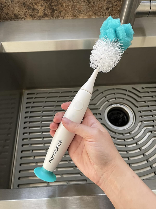 A hand holding a cleaning brush with a white bristle head and blue sponge, placed next to a stainless steel sink.