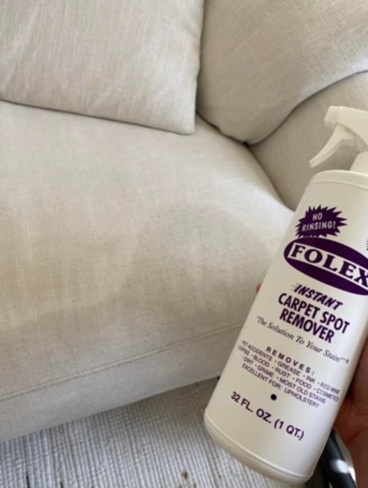A hand holding a bottle of Folex Instant Carpet Spot Remover near a sofa, indicating its use for upholstery cleaning