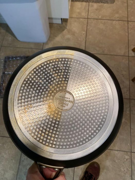 An image of the bottom of a non-stick frying pan with a stainless steel induction base, held over a tiled floor