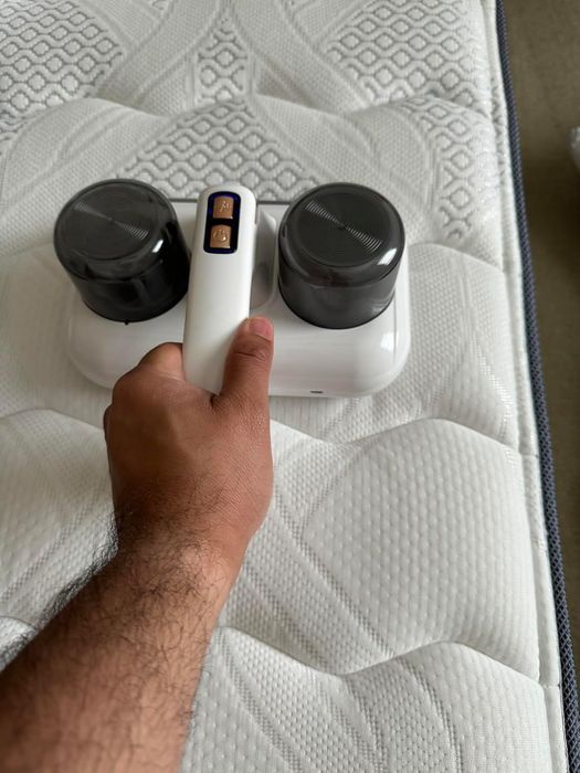 A hand holding a modern handheld vacuum designed for mattress cleaning, featuring two circular components. The image is shown in a shopping context.
