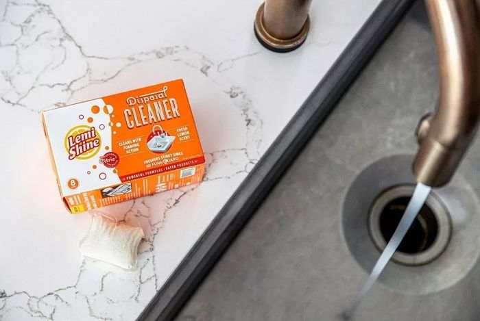 A box of Lemi Shine Disposal Cleaner placed on a kitchen countertop near a faucet and sink drain.