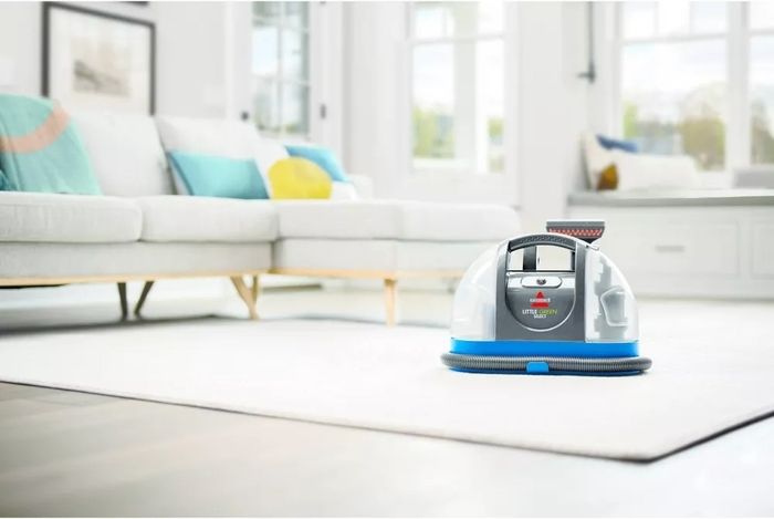 A compact Bissell Little Green ProHeat carpet cleaner is placed on a white rug in a modern living room with a sofa in the background