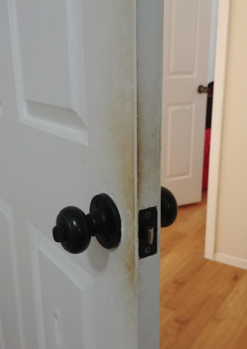 A partially open white door revealing a worn and dirty edge near the handle, with a wooden-floored room visible through the gap.