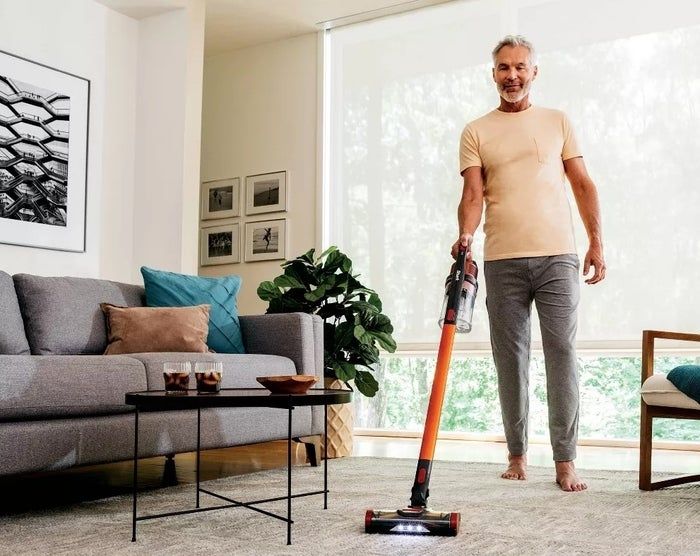 An individual using a cordless vacuum on a carpeted floor in a sleek, modern living room featuring a gray sofa, coffee table, and an indoor plant.