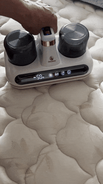 A hand using a handheld vacuum cleaner on a mattress. The device features multiple buttons and two cylindrical compartments.