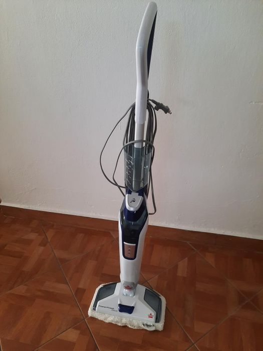 A reviewer’s upright steam mop with a detachable handheld steamer, corded, resting on a tiled floor.