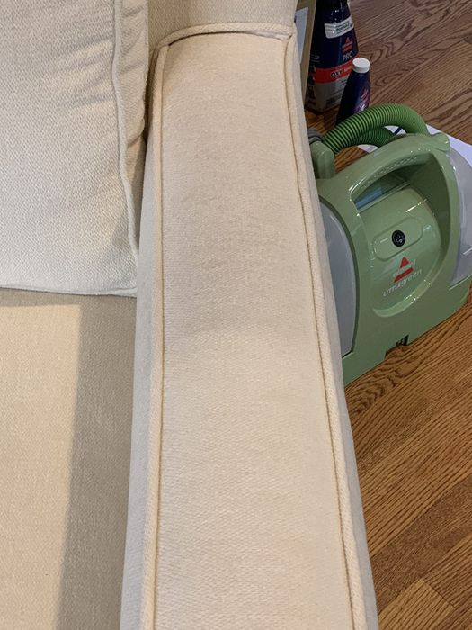 A partial view of a beige upholstered chair arm with a vertical stain and a green Bissell Little Green ProHeat cleaner placed on a wooden floor