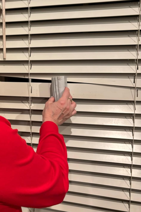 A person wearing a red shirt using a brush to clean dust from window blinds
