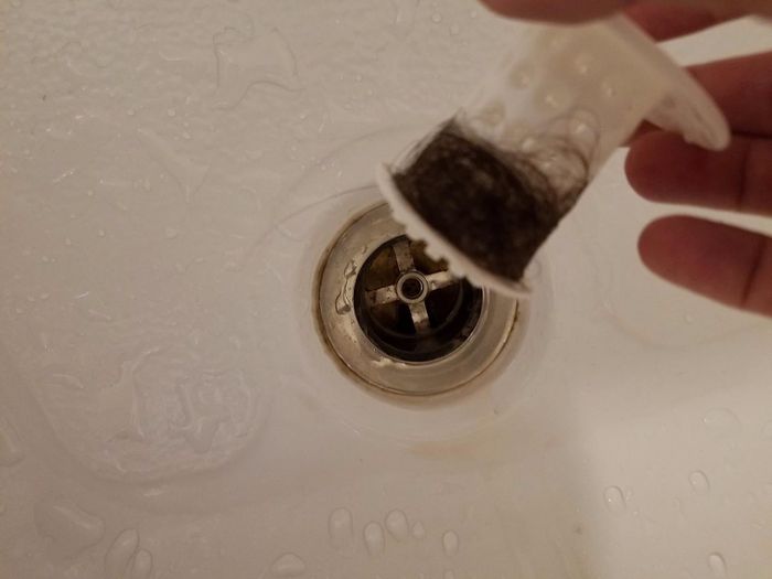 A reviewer’s hand holds a white drain hair catcher filled with hair, positioned over a bathtub drain.