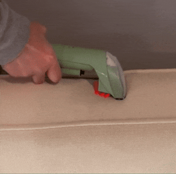 A hand using a green handheld vacuum cleaner to clean a beige sofa cushion