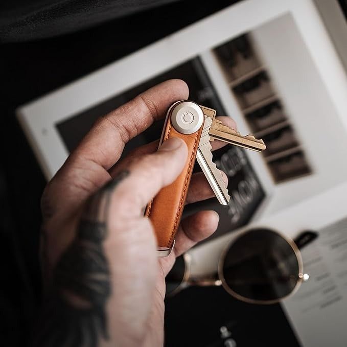 A hand holding a compact leather key organizer with keys. Below it, a tablet and sunglasses are placed on a surface.