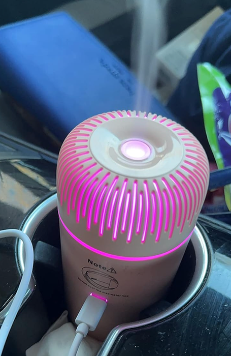 A small, rose gold cylinder-shaped diffuser, glowing pink and plugged into a drink holder