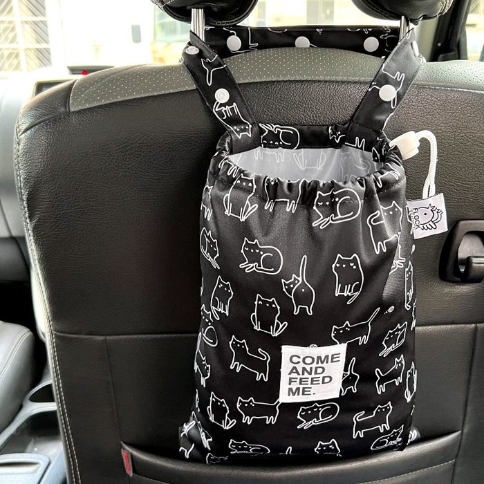 A black hanging bag with a white outline featuring a cat illustrated pattern, suspended from the back of a car seat