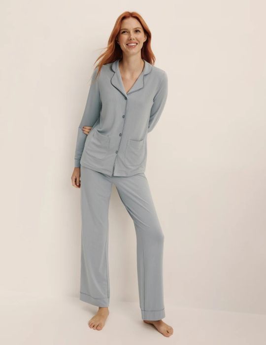 A person smiles while wearing a ribbed pajama set with long sleeves and pants, standing barefoot against a neutral background. Ideal for a shopping feature.