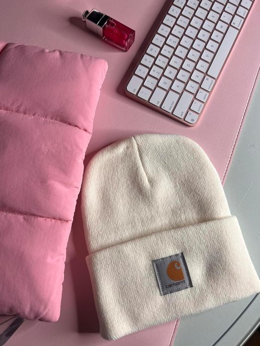 A Carhartt beanie rests on a soft pink surface, surrounded by a keyboard, pink lipstick, and a quilted jacket. The image is perfect for a shopping feature.