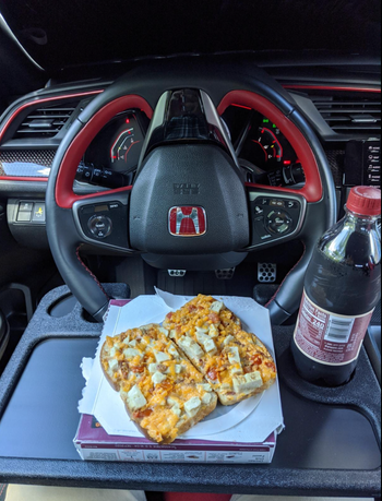A black plastic floating tray mounted on a steering wheel, with a sandwich and a coke placed on it