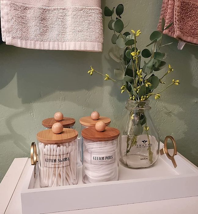 A reviewer photo of labeled glass jars with wooden lids, filled with cotton swabs and pads, placed on a tray alongside a vase of eucalyptus