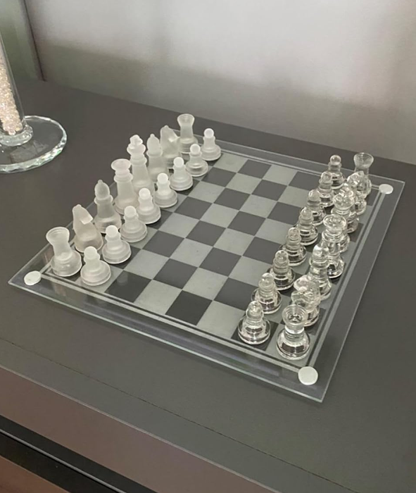 A clear glass chess set neatly arranged for the start of a game on a dark table.
