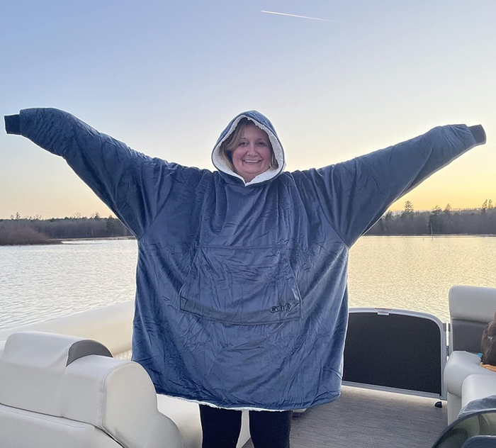 A smiling individual standing on a boat, wearing an oversized hooded blanket, arms spread wide, embracing the freedom of the open sea.