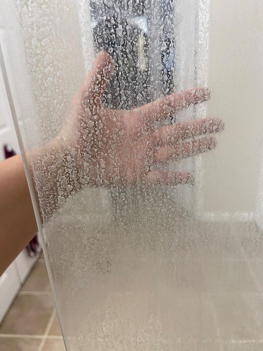 A reviewer's hand partially obscured behind a glass shower door covered in cloudy spots