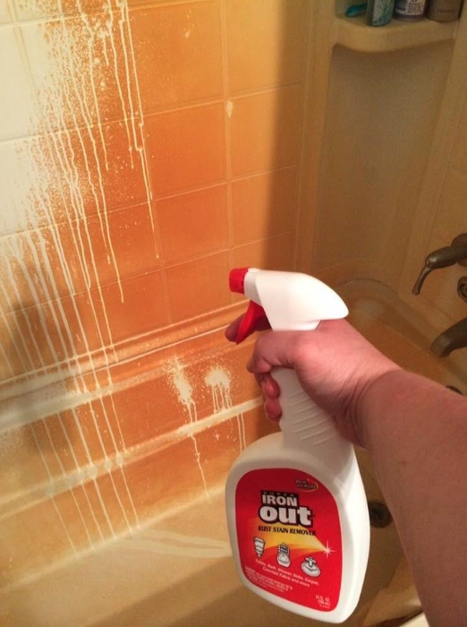 A reviewer’s hand spraying a product on a shower wall completely stained orange with rust; the rust disappears where the product has been applied.