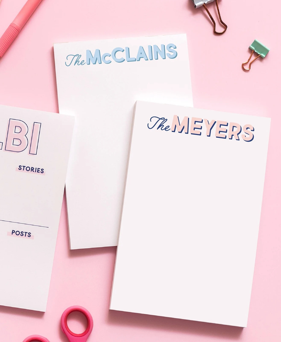 Two personalized notepads surrounded by office supplies, set against a soft pink background.