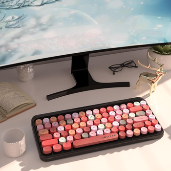 A vibrant pink and orange mechanical keyboard with round keys sits on a desk next to a monitor, an open book, a mug, a plant, and a decorative deer figurine