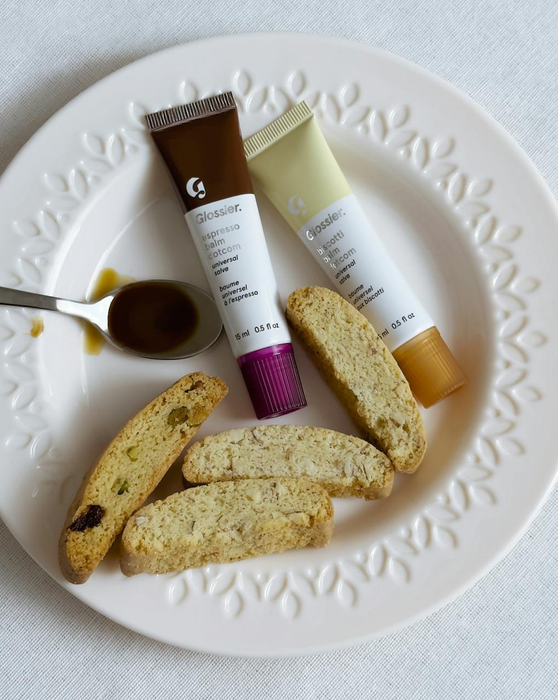 Two Glossier lip balm tubes set on a plate alongside biscotti cookies and a small spoon of honey
