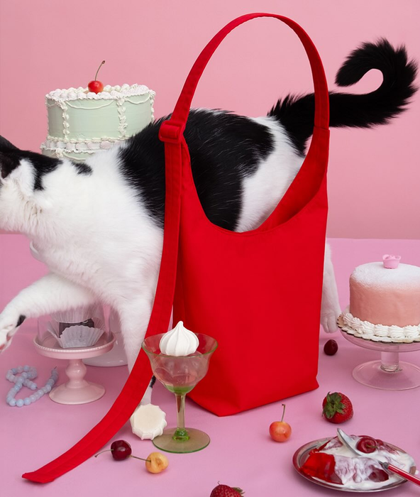 A cat sitting next to a red handbag on a table filled with cakes, fruits, and a spilled dessert, suggesting a focus on fashion accessories in a shopping context.