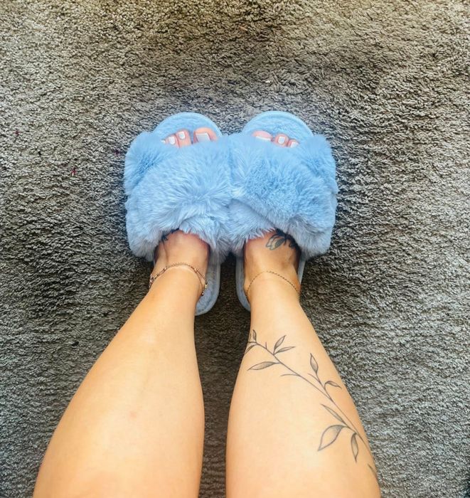 A person wearing fuzzy slippers, showing off cozy footwear on a textured surface, with tattoos visible on their legs