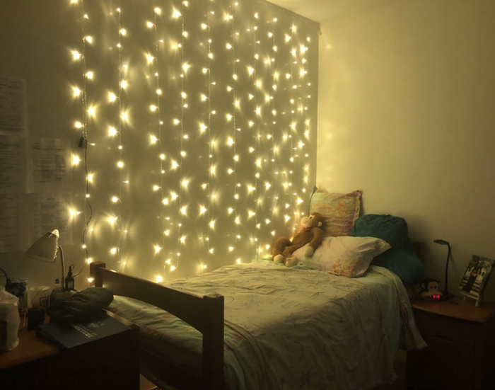 reviewer's yellow-tinted string light curtain hanging behind their bed
