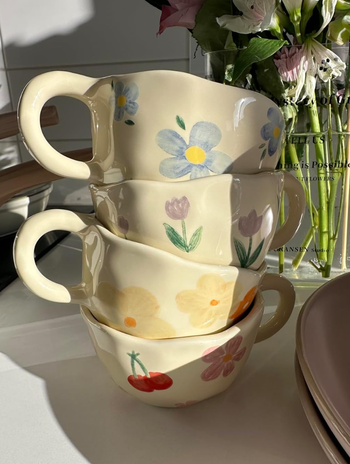 Four ceramic mugs featuring hand-painted floral and cherry designs, stacked on a white surface with fresh flowers in the background