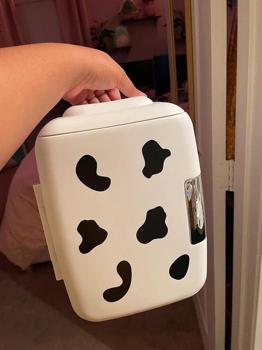 An individual holding a small, cow-patterned mini fridge with a clear front window, ideal for storing beauty and skincare products.