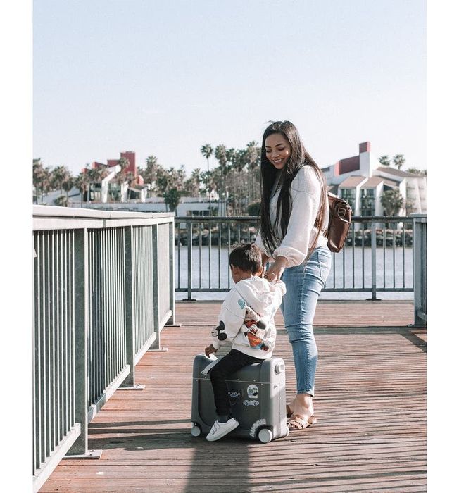 A child model sitting on the wheeled suitcase while an adult model pulls them through the airport.