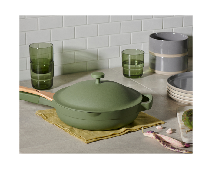 The green Always Pan sits on a kitchen counter, surrounded by cups, bowls, and plates.