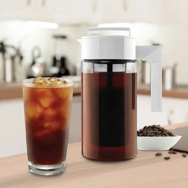 A white cold brew coffee maker placed beside a glass filled with iced coffee.