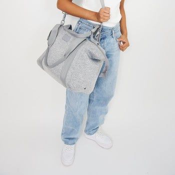Model carrying the Dagne Dover duffle bag on their shoulder, with the long shoulder strap and a zipper closure across the top, in heather grey.