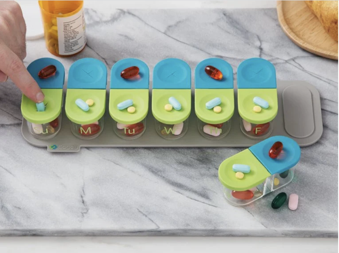 A set of seven clear pill containers with blue and green silicone lids marking day and night, placed on a gray magnetic holder