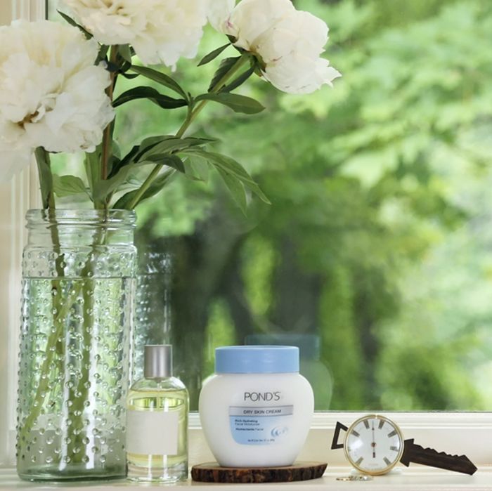 A jar of facial moisturizer, a timepiece, a fragrant room spray, and a decorative vase filled with blooming flowers.