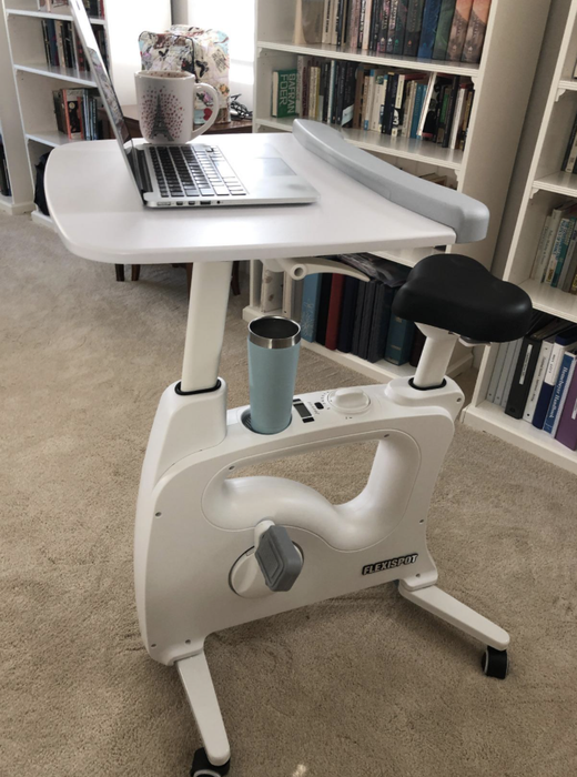 A reviewer shows a white bike desk set up in a home, with a laptop on the tray and a drink in the holder.