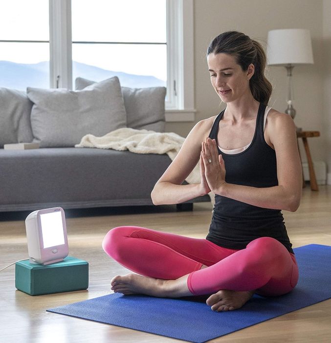 The model is seated on a yoga mat beside a small light therapy lamp.