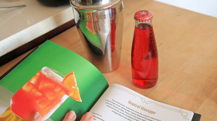 A cookbook lies open on a counter, featuring a small red SanBitter in the background alongside a metal shaker, setting the scene for a creative mixology session.