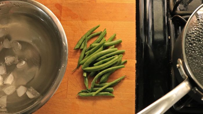 A bowl filled with ice water placed next to fresh, trimmed green beans and a pot of boiling water.