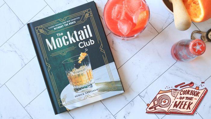 A vibrant mocktail cookbook placed on a kitchen counter, surrounded by freshly juiced oranges and a glass of red mocktail.