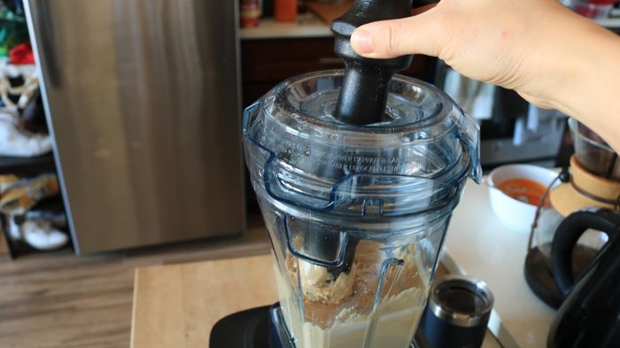 A person using a plastic tamper to press the nut butter inside a blender.