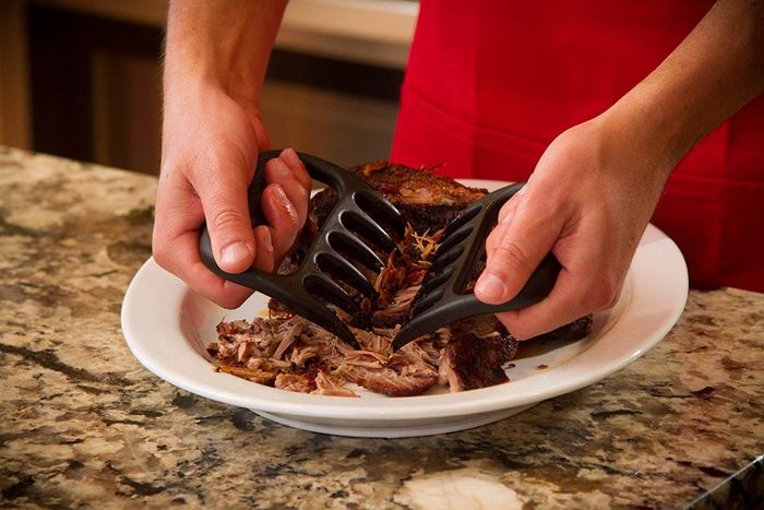 A model is using the black, claw-shaped shredders to shred meat on a plate.