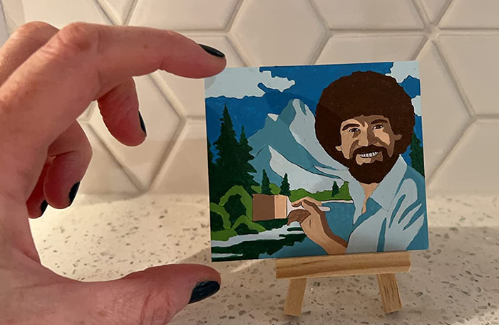 A reviewer pinching their tiny painting — a Bob Ross landscape — to show that it's smaller than their hand.